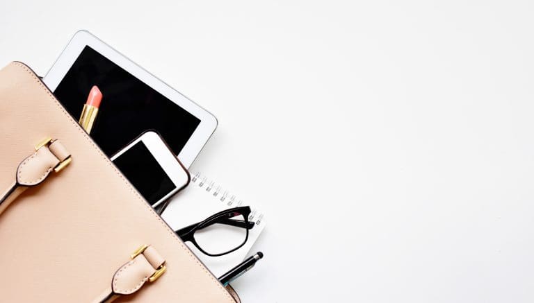 Flat lay of a blush handbag with a tablet, smartphone, lipstick, glasses and notebook on white