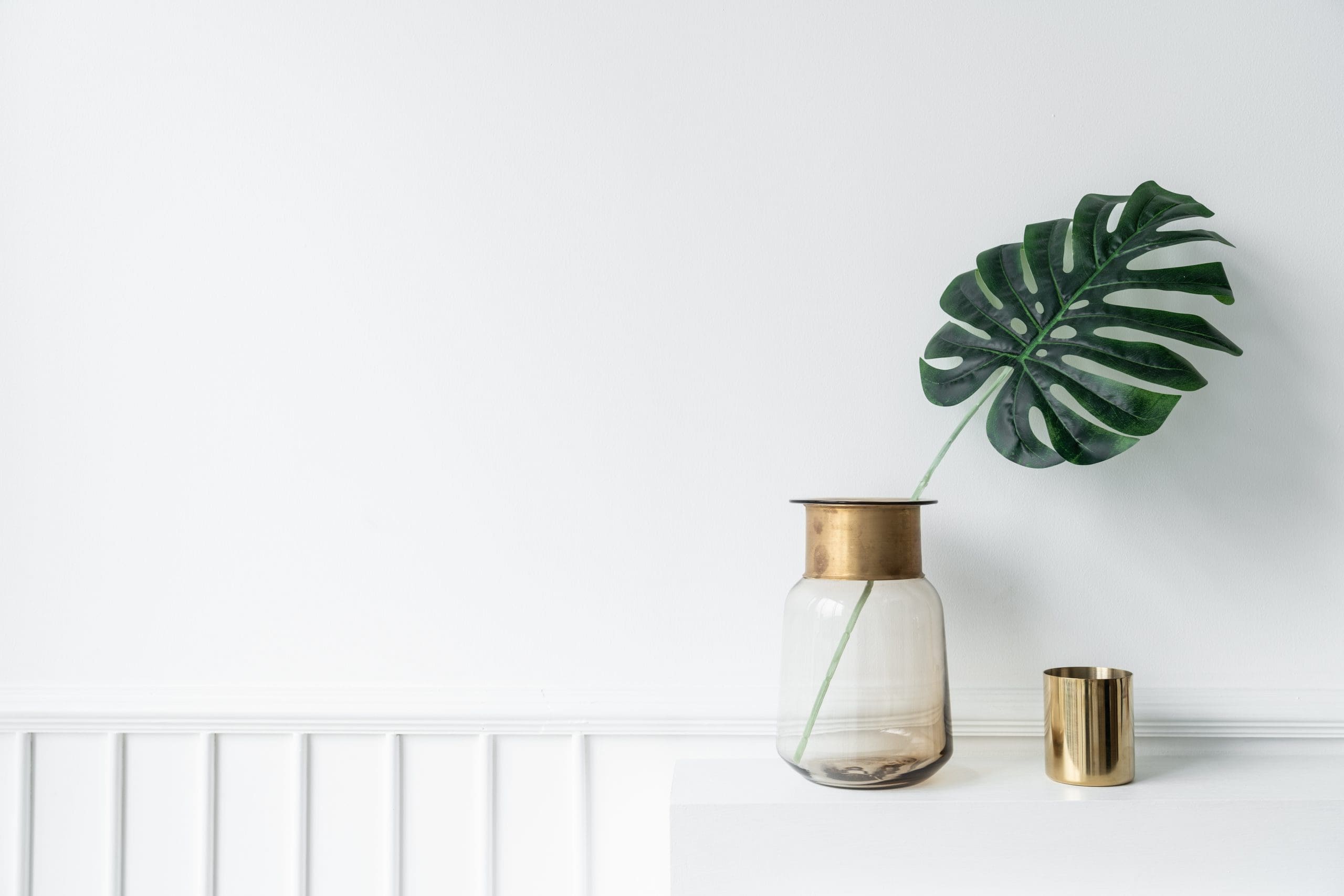 Monstera leaf in a glass vase beside a small gold cup on a white shelf