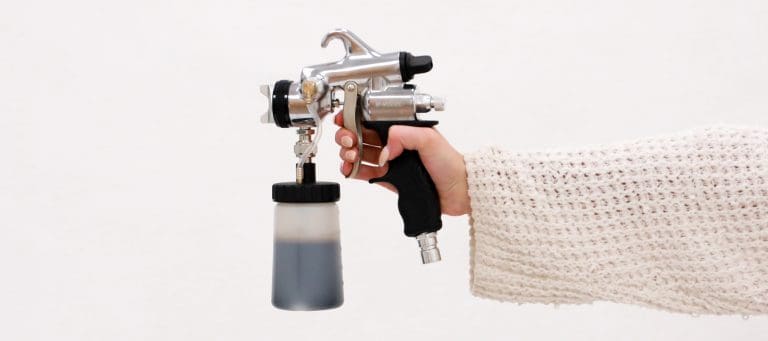 Hand in a knit sleeve holding an HVLP spray tan gun with a solution cup