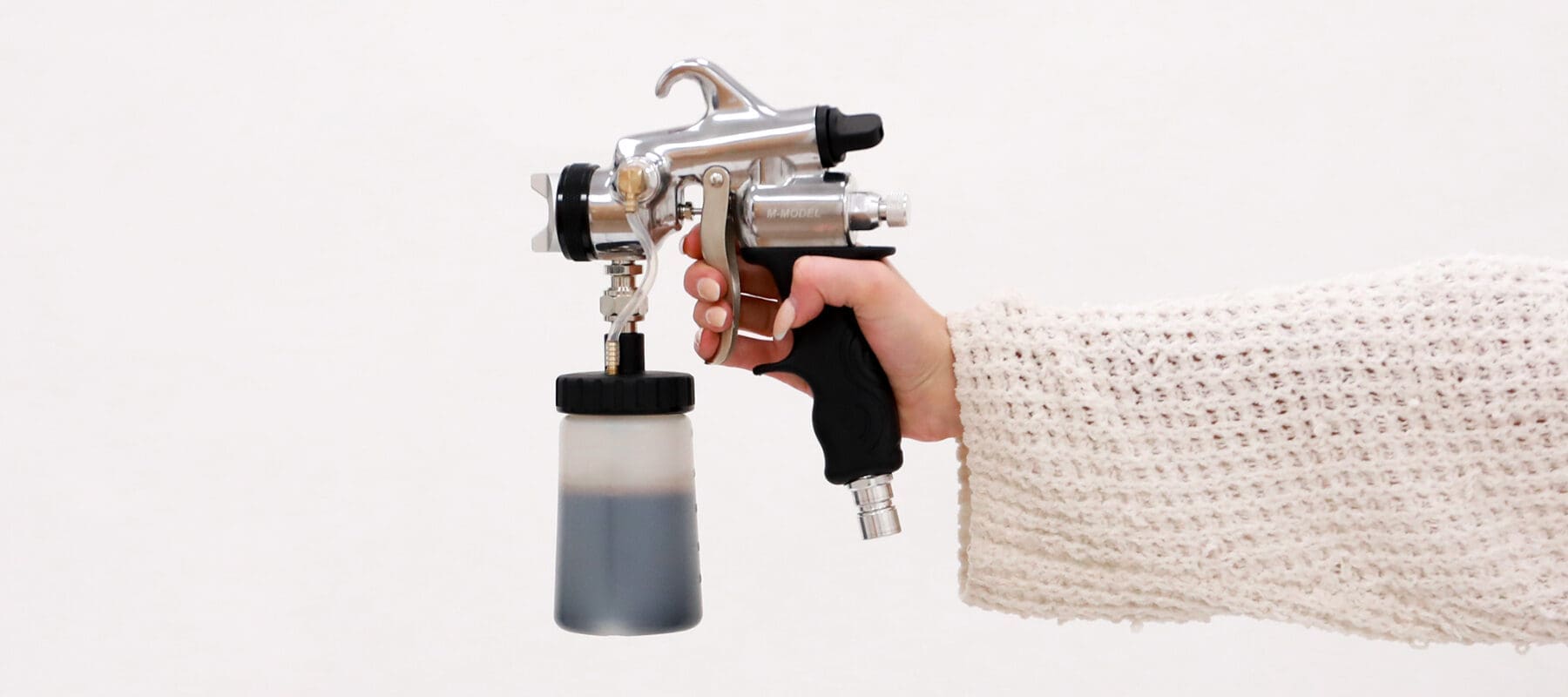 Hand in a knit sleeve holding an HVLP spray tan gun with a solution cup