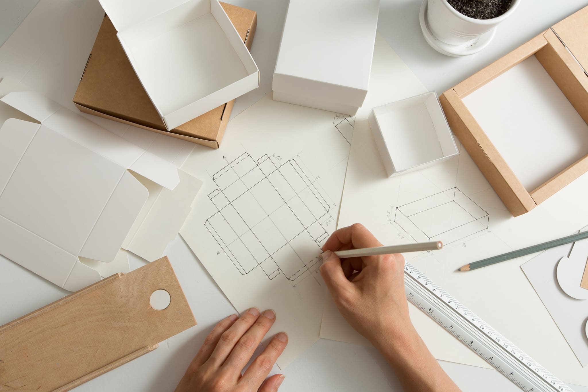 Hands sketching a box template on paper among kraft and white packaging boxes
