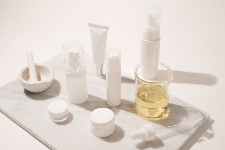 Blank white skincare bottles, jars and a dropper beside a beaker of golden oil and a mortar on a marble tray