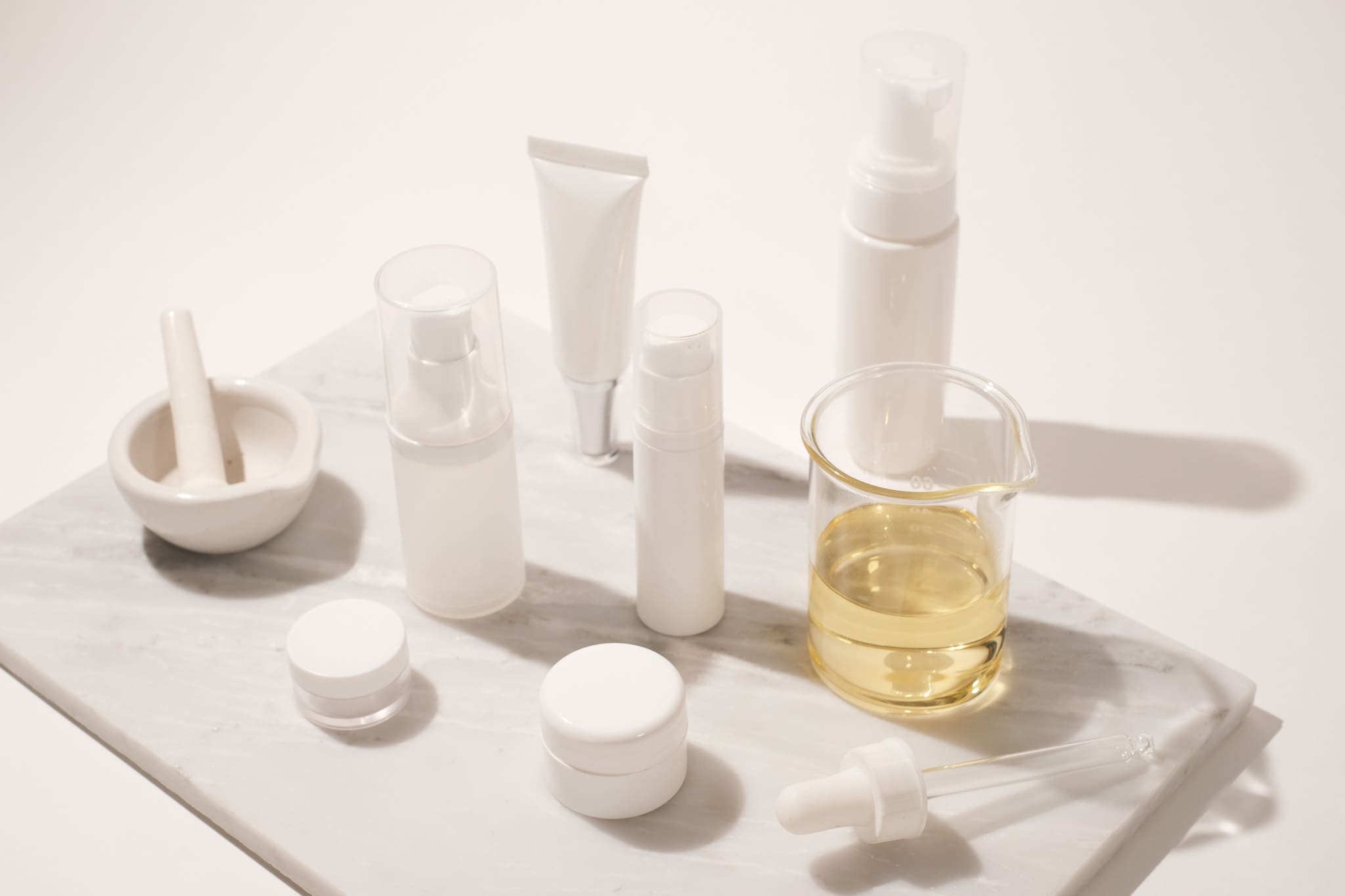 Blank white skincare bottles, jars and a dropper beside a beaker of golden oil and a mortar on a marble tray