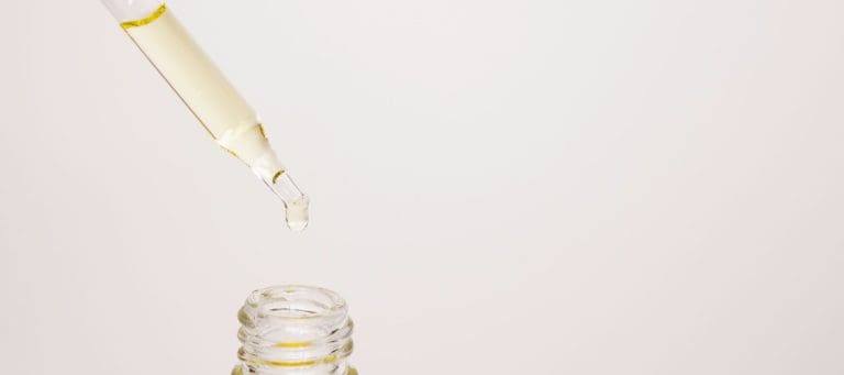 Glass dropper releasing a drop of golden oil into a small bottle on a blush background