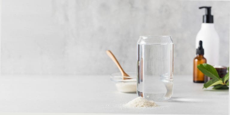Glass of water, a bowl of collagen powder with a wooden spoon, and skincare bottles beside green leaves