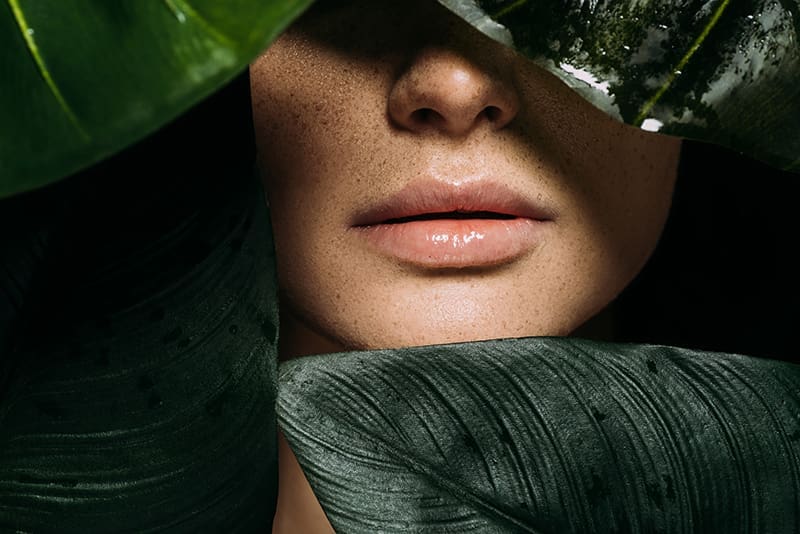 Close-up of a woman's freckled face, nose and lips framed by dark green tropical leaves