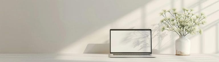 Open laptop on a white desk beside a vase of small white flowers in soft sunlight